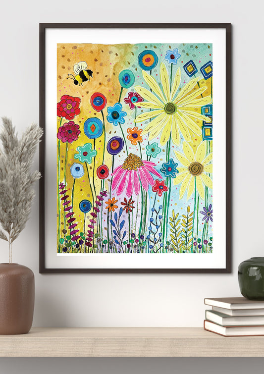 A framed art print featuring a colorful, whimsical watercolor-style wildflower scene with a bee, displayed on a shelf in a styled room setting.