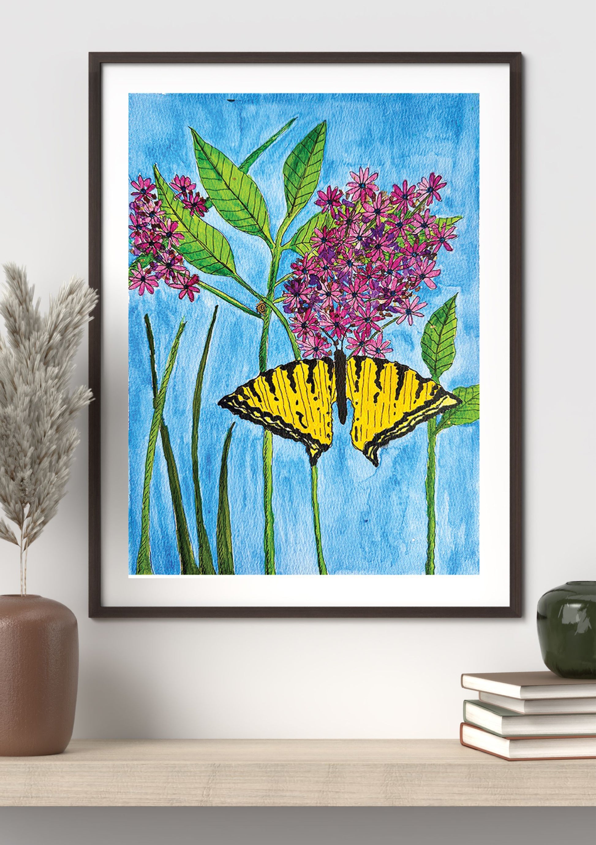 A framed art print featuring a yellow and black swallowtail butterfly perched on pink-purple flowers with green leaves against a bright blue background, displayed on a shelf in a styled interior setting.