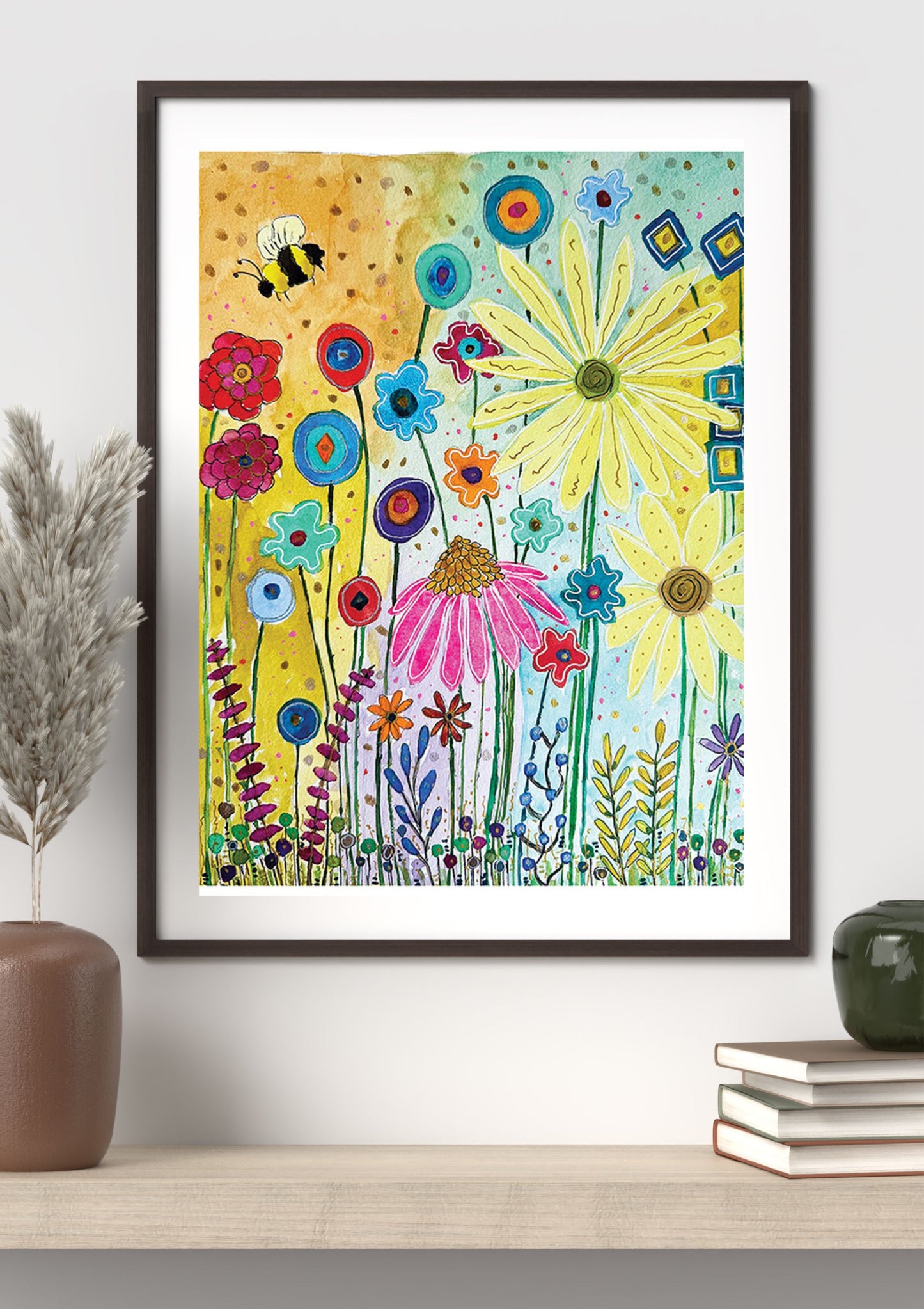 A framed art print featuring a colorful, whimsical watercolor-style wildflower scene with a bee, displayed on a shelf in a styled room setting.