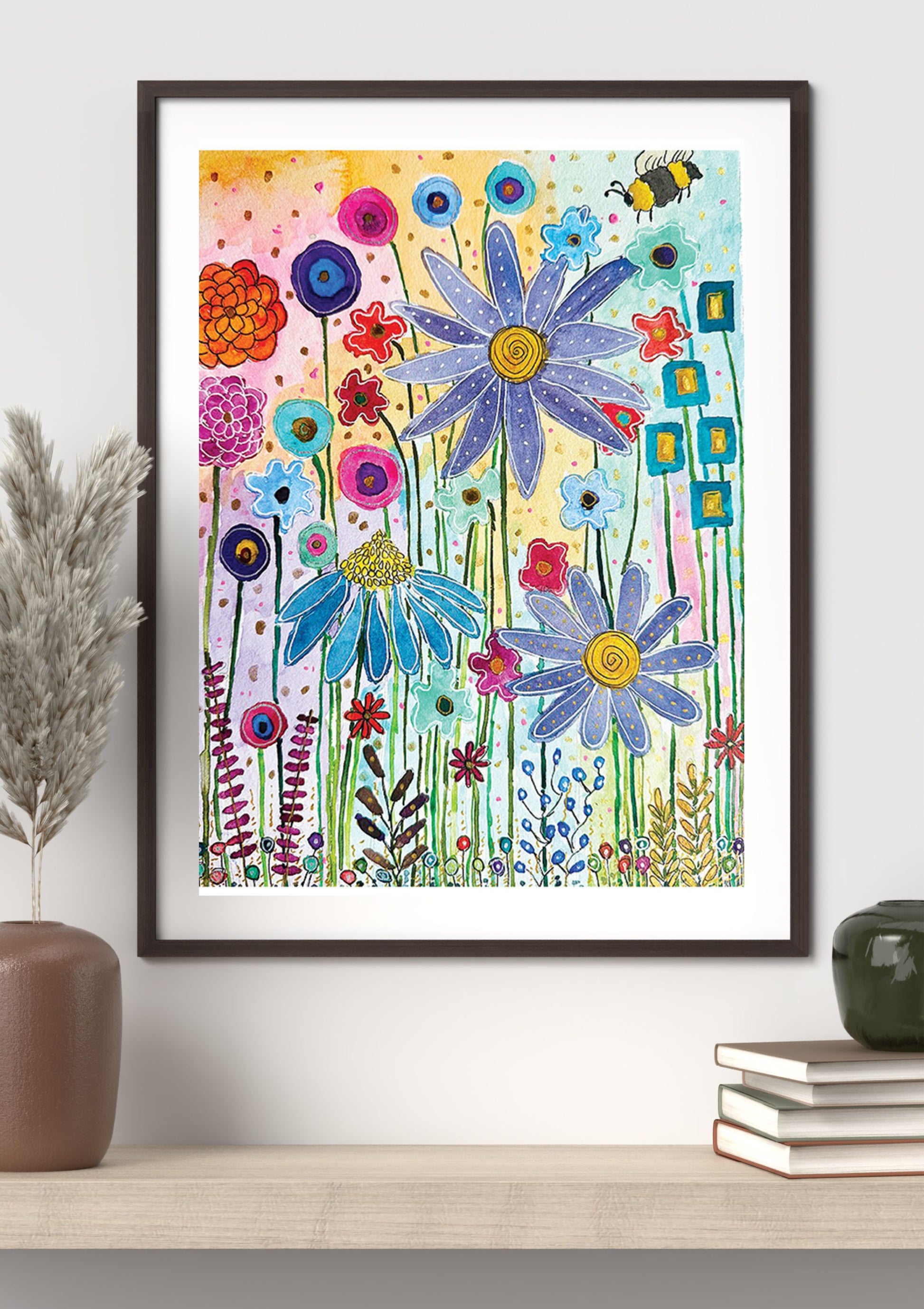 A framed colorful floral art print hangs on a white wall above a light wood shelf, styled with a brown vase on the left and stacked books on the right.