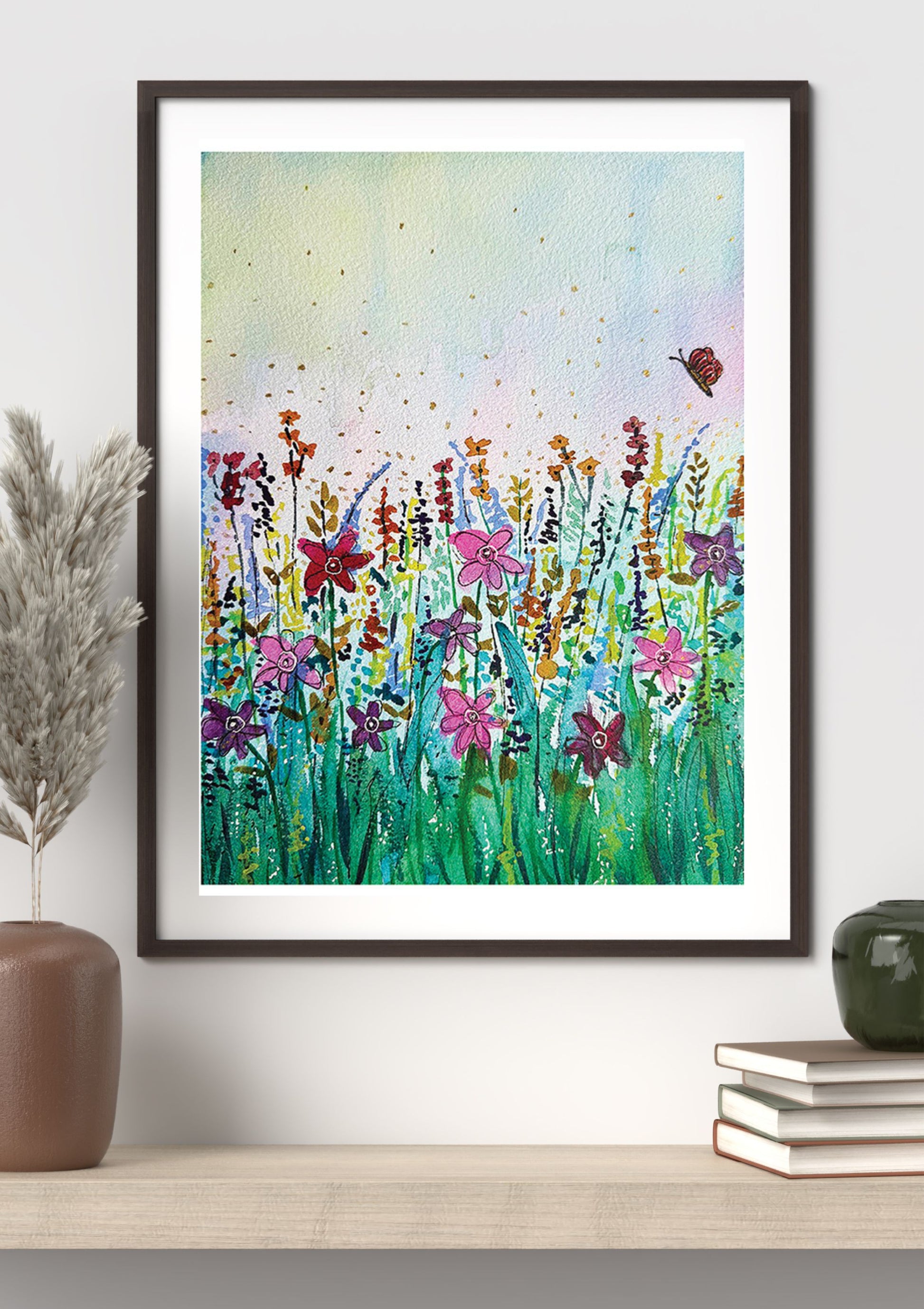 A framed watercolor art print depicting a vibrant wildflower meadow with a butterfly, styled on a light wood shelf next to a brown vase and books against a white wall.